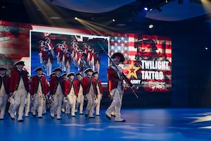 Army Soldiers dressed in red, Revolutionary War-era uniforms are marching in formation during an indoor show. The words "U.S. Army Twilight Tattoo" and other graphics appear on the screens in the background.