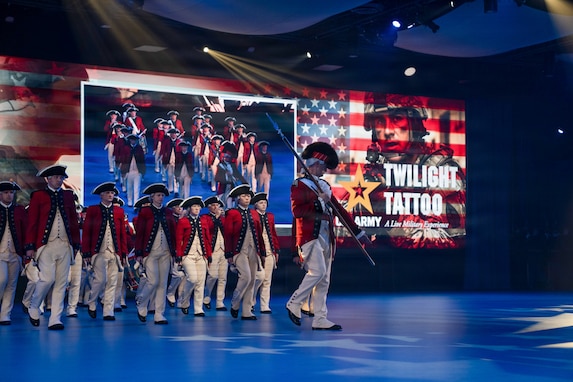 Army Soldiers dressed in red, Revolutionary War-era uniforms are marching in formation during an indoor show. The words "U.S. Army Twilight Tattoo" and other graphics appear on the screens in the background.