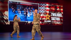 Two Army Soldiers in combat uniform are walking around while singing into microphones during an indoor show. The words "U.S. Army Twilight Tattoo" and other graphics are visible on the screen behind them.