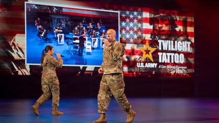 Two Army Soldiers in combat uniform are walking around while singing into microphones during an indoor show. The words "U.S. Army Twilight Tattoo" and other graphics are visible on the screen behind them.