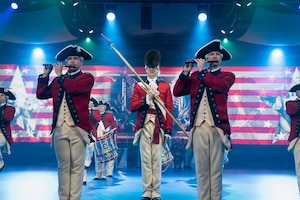 Soldiers dressed in red, Revolutionary War-era uniforms are performing on fifes and drums during an indoor performance. There are several spotlights in the background, and red and white stripes and other images projecting on a screen on the back wall.