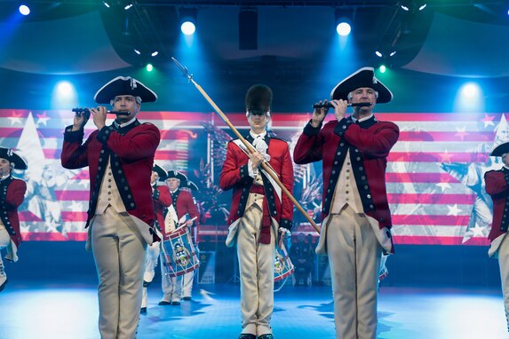 Soldiers dressed in red, Revolutionary War-era uniforms are performing on fifes and drums during an indoor performance. There are several spotlights in the background, and red and white stripes and other images projecting on a screen on the back wall.