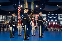 Army Soldiers in various ceremonial and period uniforms are marching in formations across a blue-colored floor at an indoor event.