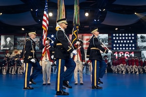 Army Soldiers in various ceremonial and period uniforms are marching in formations across a blue-colored floor at an indoor event.