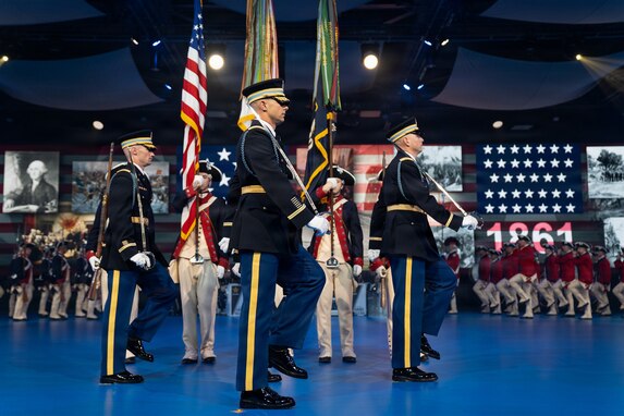 Army Soldiers in various ceremonial and period uniforms are marching in formations across a blue-colored floor at an indoor event.