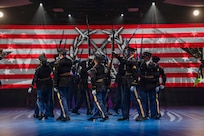 Army Soldiers dressed in dark ceremonial uniforms are marching in a circle while holding up ceremonial rifles during a drill routine at an indoor show.