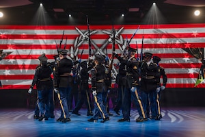 Army Soldiers dressed in dark ceremonial uniforms are marching in a circle while holding up ceremonial rifles during a drill routine at an indoor show.