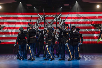 Army Soldiers dressed in dark ceremonial uniforms are marching in a circle while holding up ceremonial rifles during a drill routine at an indoor show.