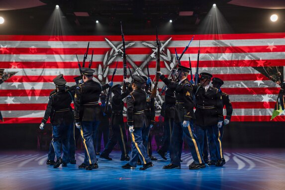 Army Soldiers dressed in dark ceremonial uniforms are marching in a circle while holding up ceremonial rifles during a drill routine at an indoor show.