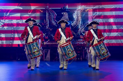 Army soldiers dressed in red Revolutionary War-era uniforms are performing on the drums during an indoor show. Behind them is a large screen projecting red and white stripes and a large emblem that includes an eagle.