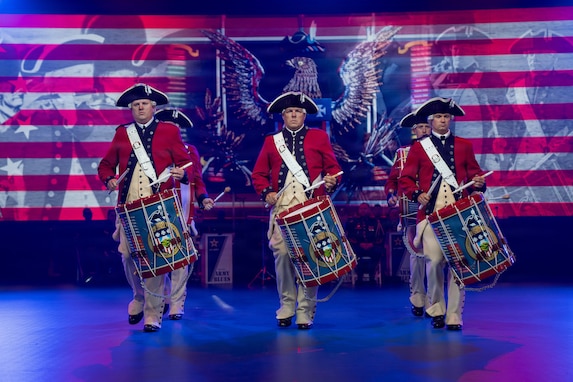 Army soldiers dressed in red Revolutionary War-era uniforms are performing on the drums during an indoor show. Behind them is a large screen projecting red and white stripes and a large emblem that includes an eagle.