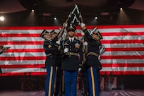 Army Soldiers dressed in dark ceremonial uniforms are performing a drill routine with ceremonial rifles during an indoor show. There is large screen in the background with red and white stripes and other images projected onto it.