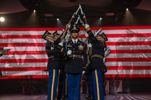 Army Soldiers dressed in dark ceremonial uniforms are performing a drill routine with ceremonial rifles during an indoor show. There is large screen in the background with red and white stripes and other images projected onto it.