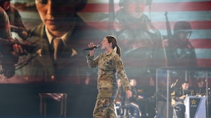 An Army Soldier dressed in combat uniform is walking across a stage during an indoor show while singing into a microphone. Images of other soldiers are projected on the wall behind her, and a military band is seated and performing in the background.