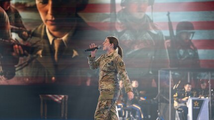 An Army Soldier dressed in combat uniform is walking across a stage during an indoor show while singing into a microphone. Images of other soldiers are projected on the wall behind her, and a military band is seated and performing in the background.
