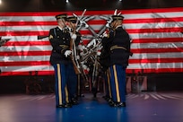 Army Soldiers dressed in dark ceremonial uniforms are standing in two rows facing each other while twirling ceremonial rifles during an indoor show. There are red and white stripes and a large insignia with a wreath surrounding two crossed rifles on the screen in the background.