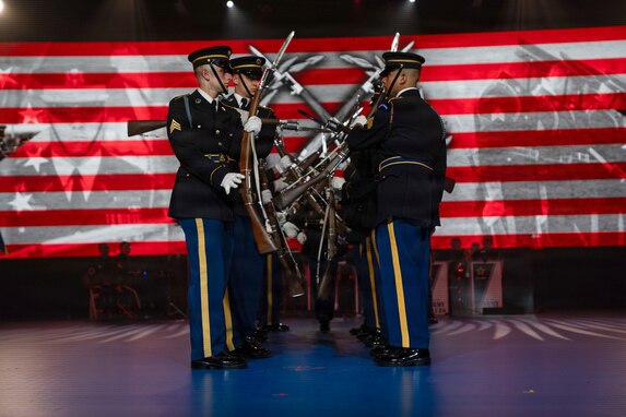 Army Soldiers dressed in dark ceremonial uniforms are standing in two rows facing each other while twirling ceremonial rifles during an indoor show. There are red and white stripes and a large insignia with a wreath surrounding two crossed rifles on the screen in the background.