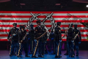 Army Soldiers dressed in dark ceremonial uniforms are marching in a circle with ceremonial rifles in their hands during an indoor show. There are red and white stripes and a large insignia with a wreath surrounding two crossed rifles on the screen in the background.