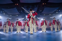Army Soldiers dressed in red Revolutionary War-era uniforms are standing in formation during an indoor event with many spotlights flashing. The soldier in front is wearing a large, furry hat and holding a long staff in front of his body while saluting.