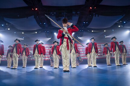 Army Soldiers dressed in red Revolutionary War-era uniforms are standing in formation during an indoor event with many spotlights flashing. The soldier in front is wearing a large, furry hat and holding a long staff in front of his body while saluting.