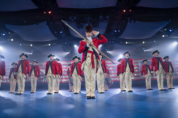 Army Soldiers dressed in red Revolutionary War-era uniforms are standing in formation during an indoor event with many spotlights flashing. The soldier in front is wearing a large, furry hat and holding a long staff in front of his body while saluting.