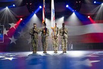 Four Army Soldiers dressed in combat uniforms are presenting the colors at an indoor event with many spotlights in the background and stars projected onto the floor.