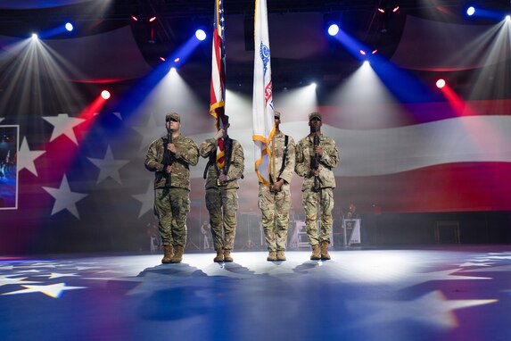 Four Army Soldiers dressed in combat uniforms are presenting the colors at an indoor event with many spotlights in the background and stars projected onto the floor.