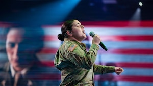 An Army soldier dressed in combat uniform sings while holding a microphone. There is an image of another soldier and U.S. Flag red and white stripes projected on the screen in the background.