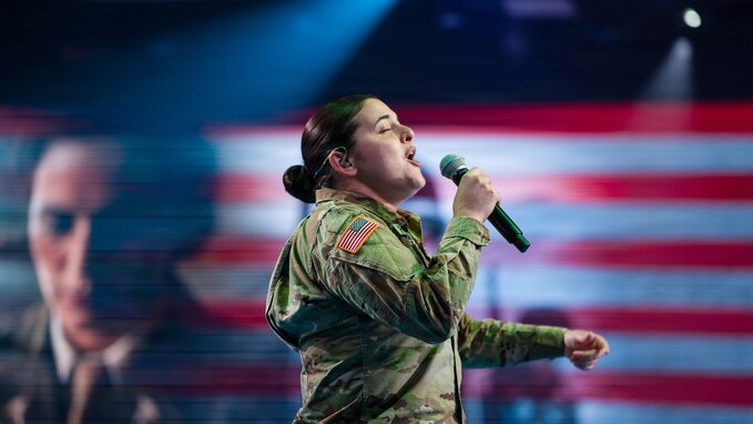 An Army soldier dressed in combat uniform sings while holding a microphone. There is an image of another soldier and U.S. Flag red and white stripes projected on the screen in the background.