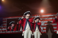Soldiers dressed in Revolutionary War-era uniforms are performing on fifes during an indoor event. There are images of the performers also projected on screens behind them.