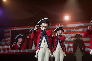 Soldiers dressed in Revolutionary War-era uniforms are performing on fifes during an indoor event. There are images of the performers also projected on screens behind them.