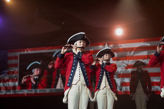 Soldiers dressed in Revolutionary War-era uniforms are performing on fifes during an indoor event. There are images of the performers also projected on screens behind them.