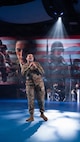 An Army soldier in combat uniform is holding a microphone and singing. There are images of soldiers and the US flag being projected on screens behind her.