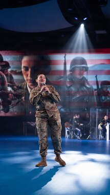 An Army soldier in combat uniform is holding a microphone and singing. There are images of soldiers and the US flag being projected on screens behind her.