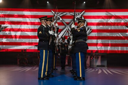 Soldiers dressed in dark ceremonial uniforms are facing each other in two rows doing a ceremonial routine with rifles at an indoor show.
