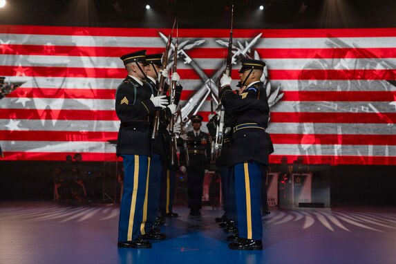 Soldiers dressed in dark ceremonial uniforms are facing each other in two rows doing a ceremonial routine with rifles at an indoor show.