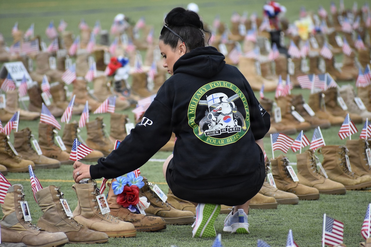 A woman wearing casual attire places a small American flag into a combat boot with the picture of a man attached to it; there are more than 7,500 combat boots lined in rows all around her on a field.