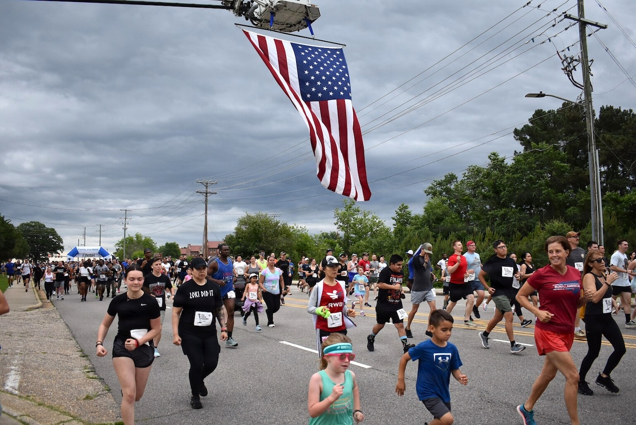 On a cloudy day, more than 2,000 people in athletic attire run down a road with a large American flag hanging overhead.