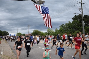 On a cloudy day, more than 2,000 people in athletic attire run down a road with a large American flag hanging overhead.