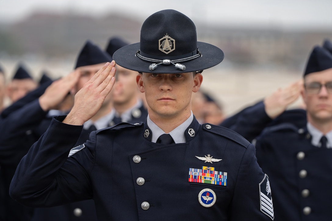 A guardian in a tall drill instructor hat salutes in front of a formation of guardians, all in dark blue dress uniforms.