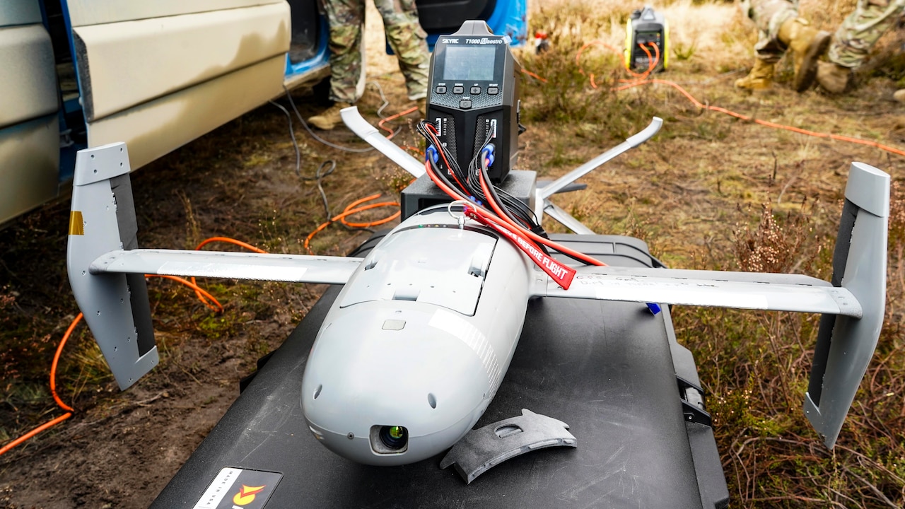 A military drone sits on the ground as military personnel stand in the background.