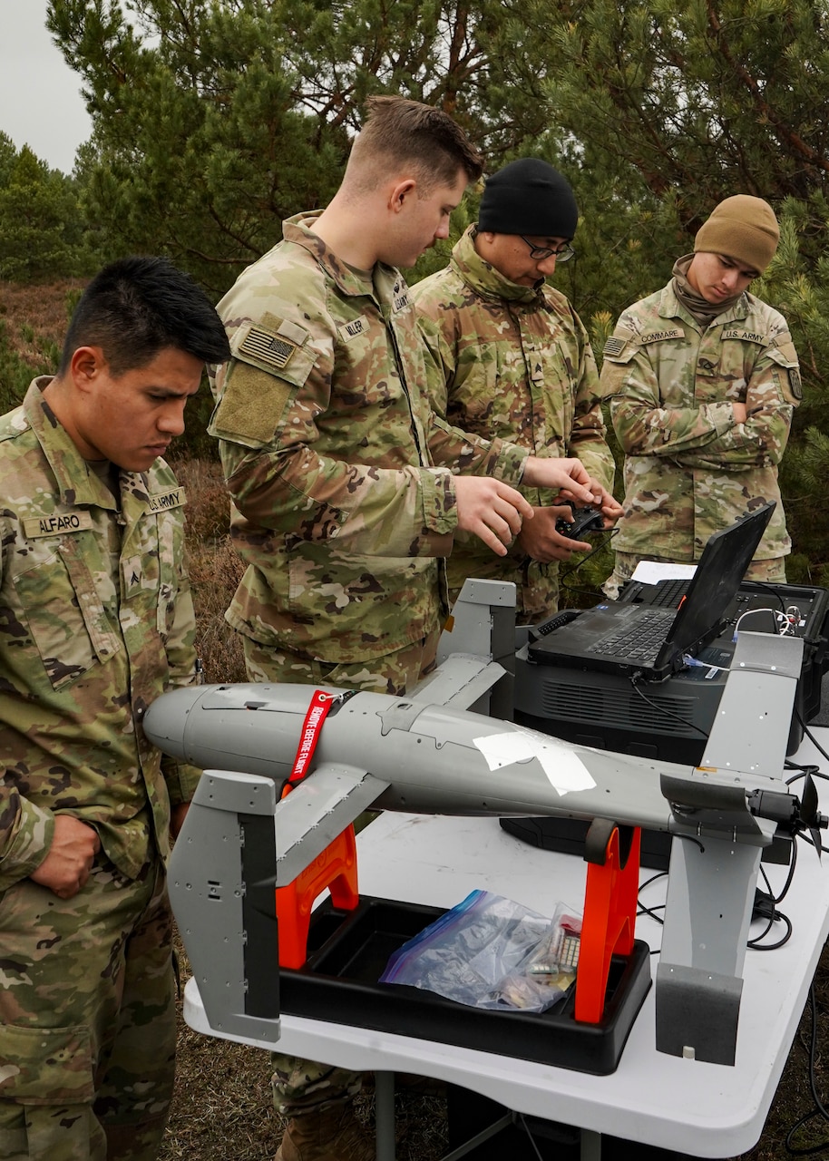 Four men wearing camouflage military uniforms look at a laptop wired to a military drone.