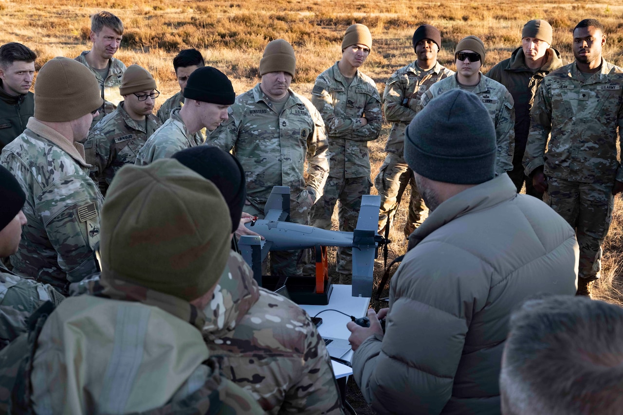 A crowd of people, some wearing camouflage military uniforms and others in winter coats, stand surrounding a military drone in an open field.