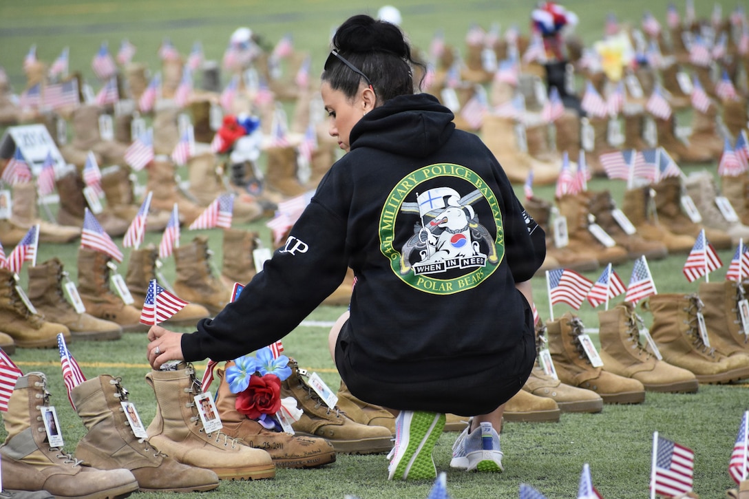 A person in running gear bends to place an American flag in one of thousands of combat boots sitting in a field during the day.