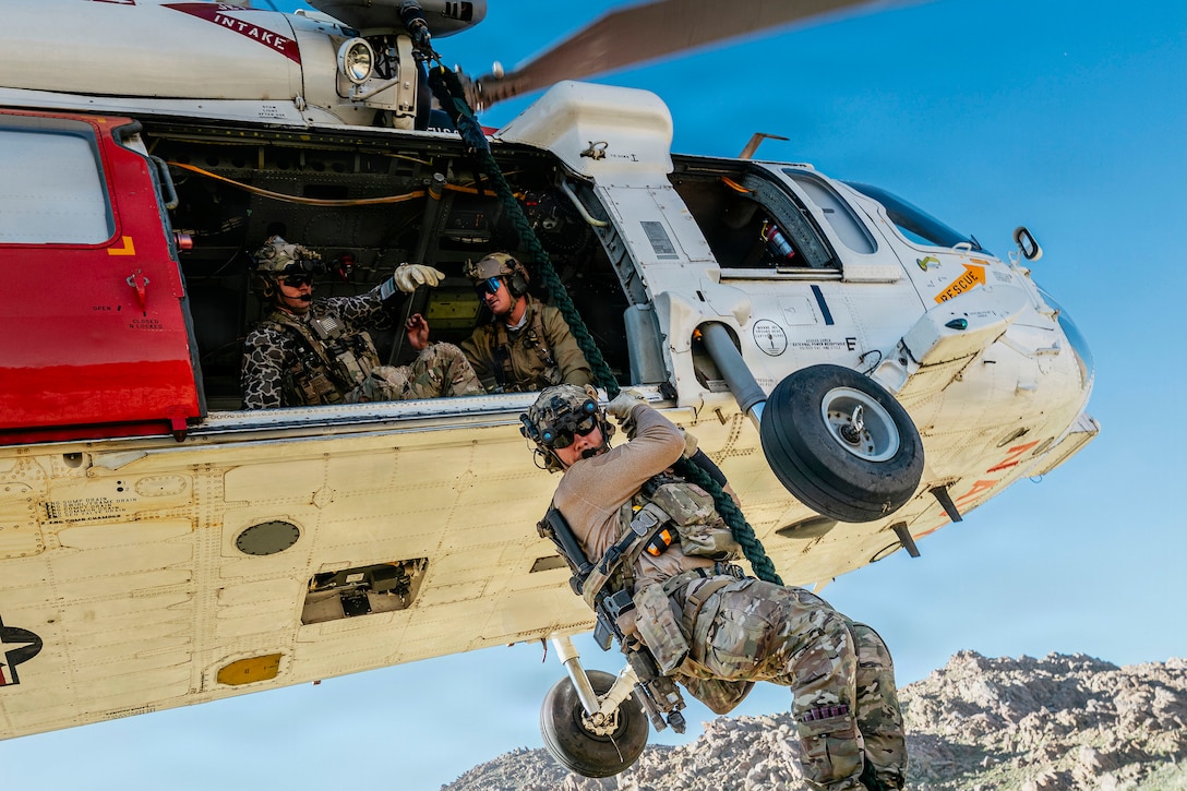 Sailors look out of the open doorway of an airborne helicopter as a fellow sailor is hoisted over grass with a mountain in the background.