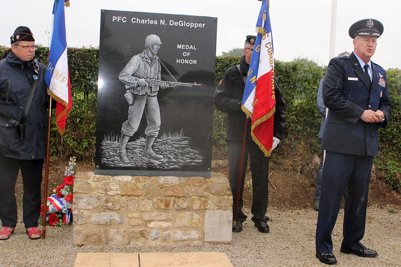A man in a formal military uniform speaks outdoors in front of a plaque featuring a soldier holding a weapon. Behind him are two men in similar attire, holding flags on each side of the plaque.