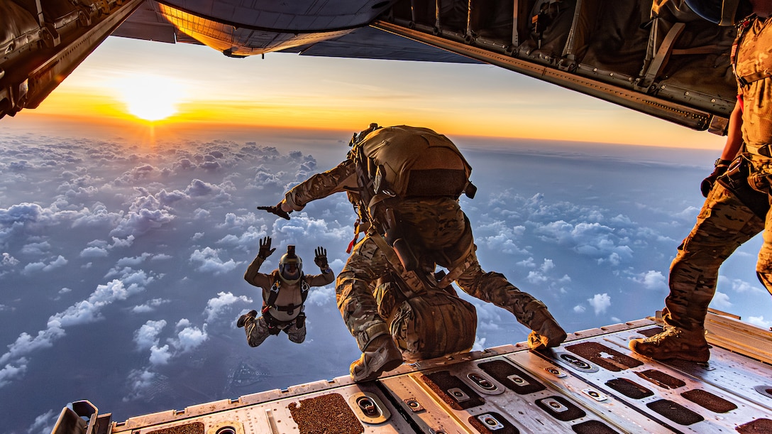 An airmen is suspended in the air by the back of an open aircraft, where another prepares to jump.