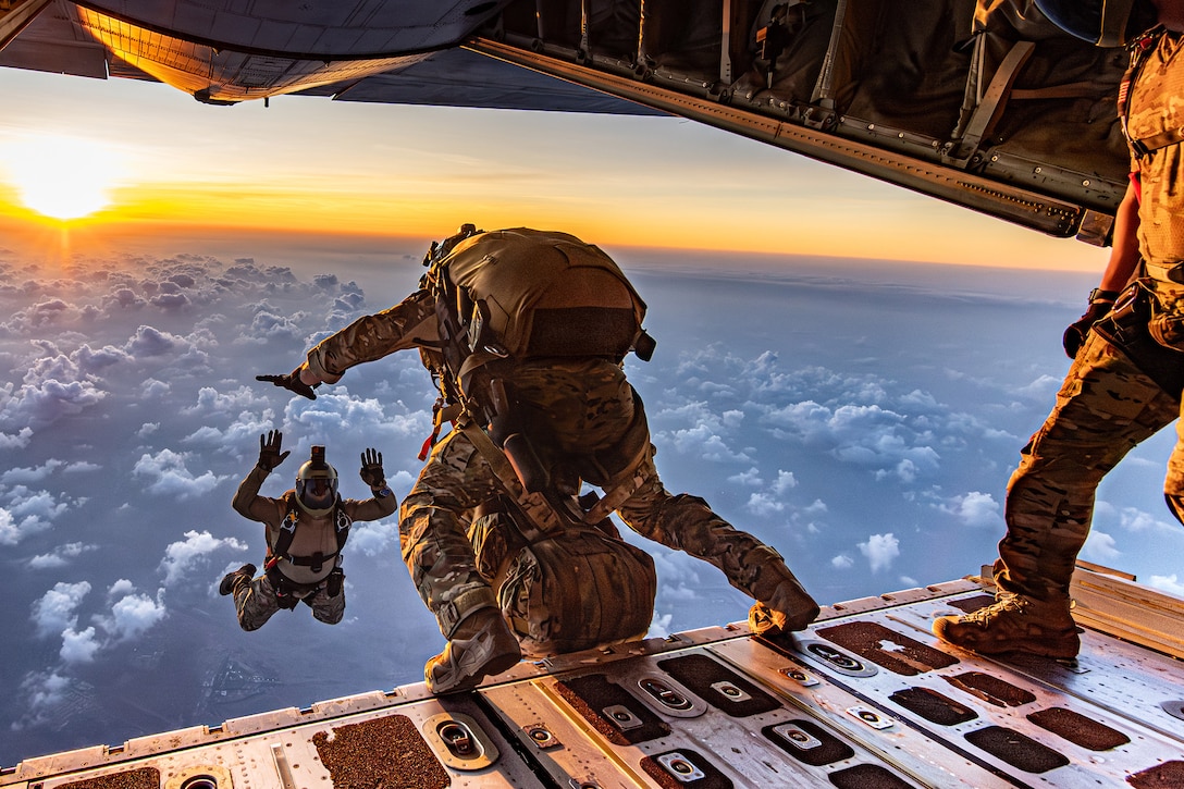 An airmen is suspended in the air by the back of an open aircraft, where another prepares to jump.