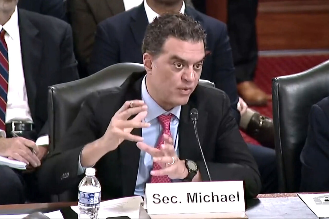 A man wearing a business suit sits at a table. In front of him is a microphone and a sign that reads "Sec. Michael."