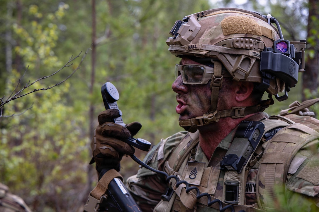 A soldier in the woods talks into a black phone receiver they are holding up in front of their face.
